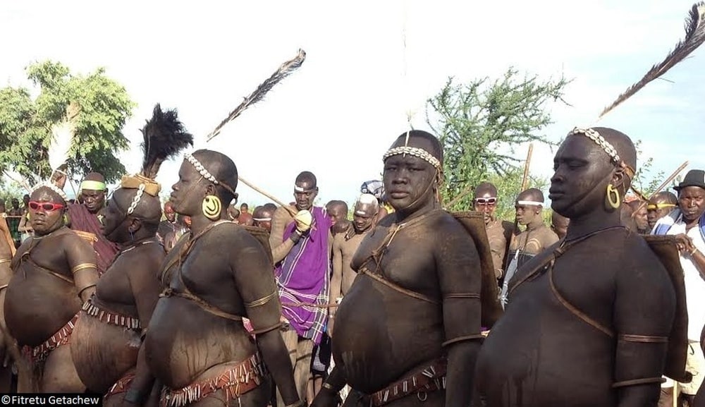 Homens da tribo perfilados exibindo suas barrigas grandes, esforçando-se para apresentar dentro do padrão de beleza.